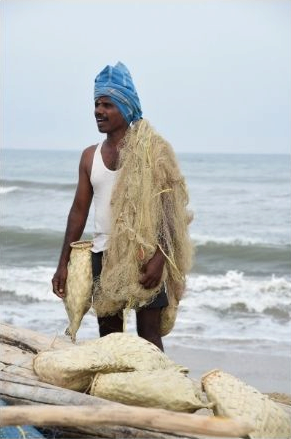 Male figure standing beside the ocean with a fishing net hanging over his left shoulder and another object in his right hand with similar items in front of him.