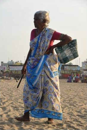 Elder woman standing on a beach in three quarter profile view. She carries a bag on one arm and a cylindrical item in her other hand.