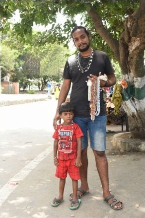 Standing male figure holding many necklaces in his left hand while putting his other hand on the shoulder of a young boy standing right up against his right leg.